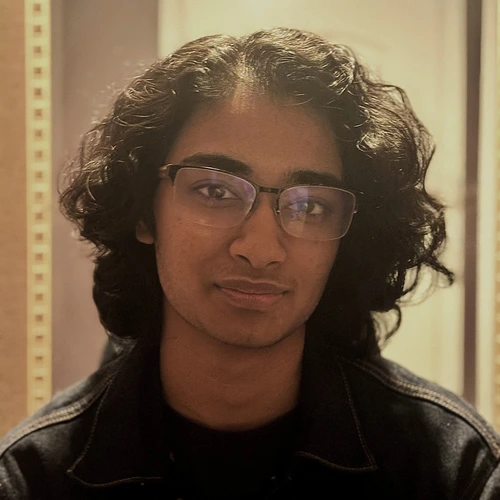 Profile picture of Jai Indra: a person with long black curly hair and wire-rimmed glasses, wearing a denim jacket, photographed indoors with warm  light.
